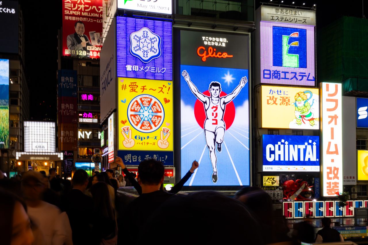 تضيء اللافتة بعد 30 دقيقة من غروب الشمس. يقلد الزوار ”وضعية غليكو“ الشهيرة: الجري بأيدي مرفوعة بعد تحقيق النصر (© Nippon.com)