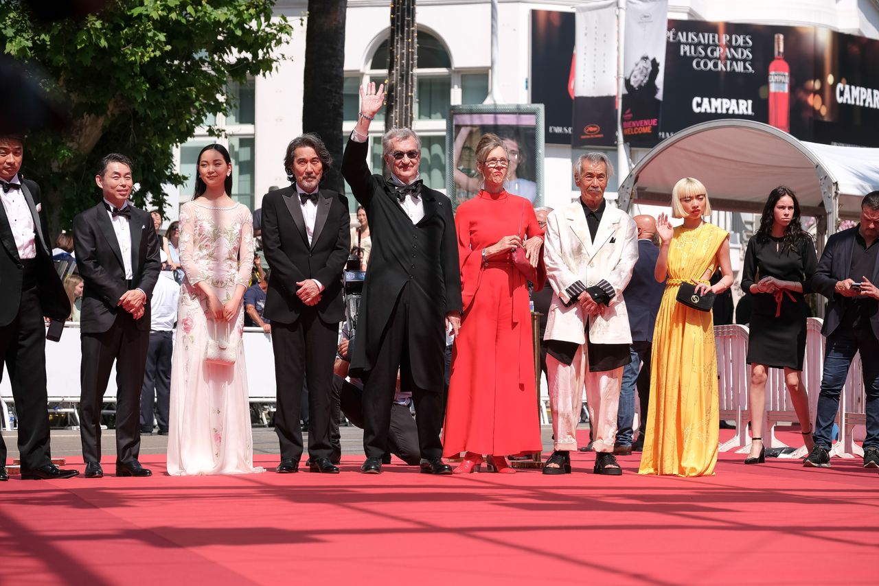 المخرج فيم فيندرز (وسط) وهو يلوح بيده للجمهور، مع الممثل ياكوشو كوجي (على جانبه اليسار) وهما يقفان على السجادة الحمراء في مهرجان كان السينمائي. الشخص الثاني من اليسار هو المنتج السيد ياناي كوجي، © واكاياما كازوكو.