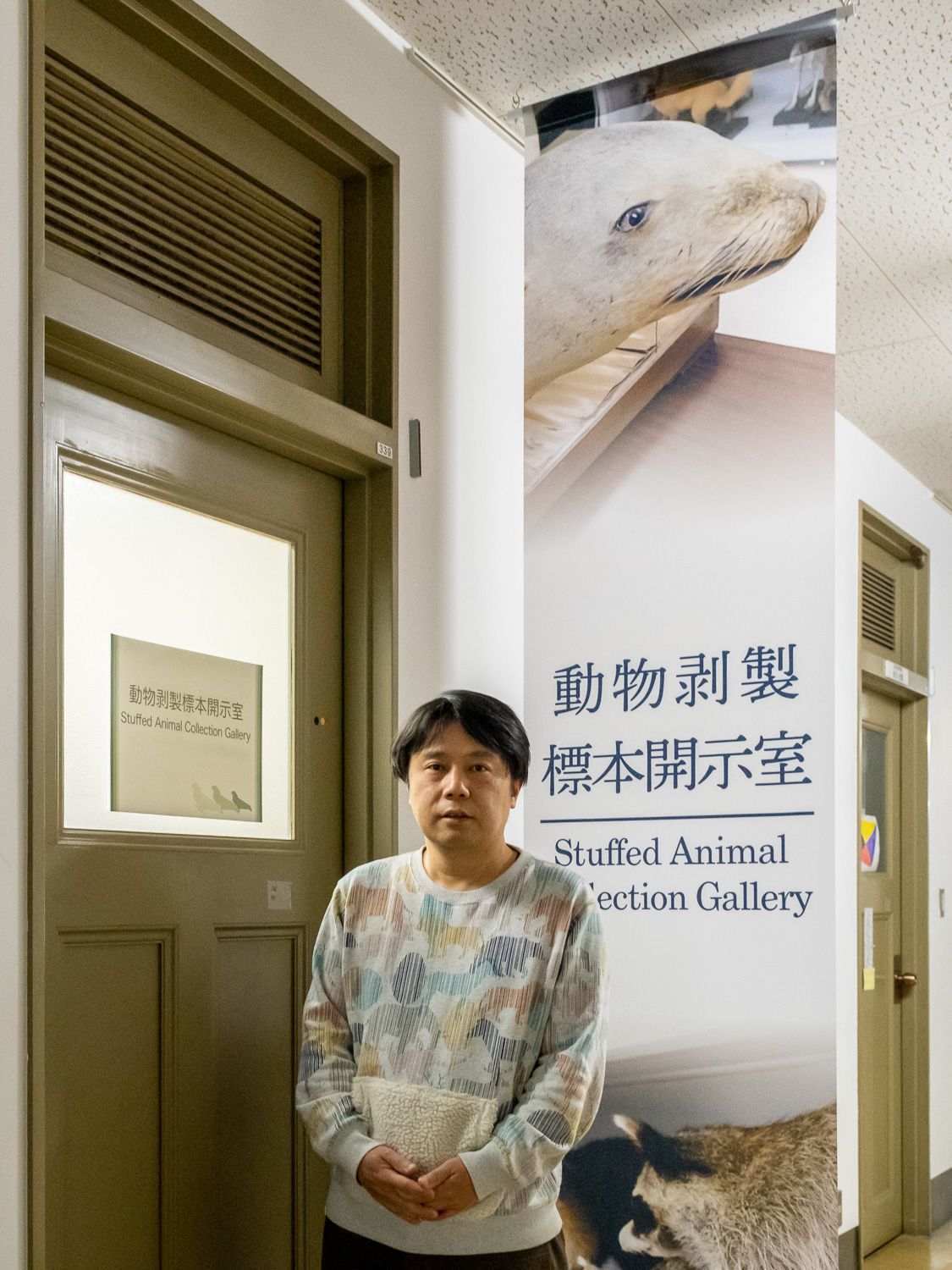 ماروياما مونيتوشي خارج معرض الحيوانات المحنطة بمتحف جامعة كيوشو. توجد لافتة معلقة في الخارج تظهر أسد البحر الياباني المحتفظ به. (© هاياشي ميتشيكو)