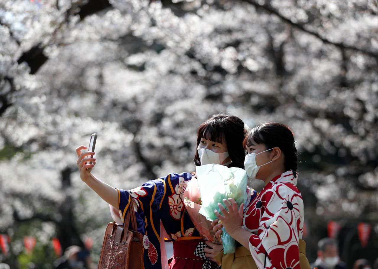فتاتان بزي الكيمونو ترتديان كمامتين واقيتين تلتقطان صور سيلفي بين أزهار الكرز المتفتحة وسط جائحة فيروس كورونا (كوفيد-19)، في طوكيو في اليابان، 23 مارس/آذار عام 2021. رويترز/إيسّيي كاتو.