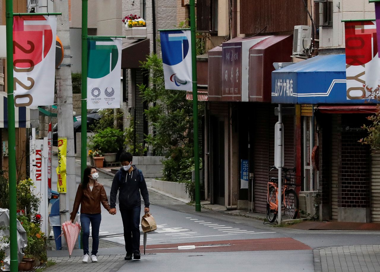 زوجان يرتديان أقنعة واقية للوجه، وسط جائحة فيروس كورونا، يشقان طريقهما في شارع تسوق محلي مزين بأعلام دورة الألعاب الأولمبية طوكيو 2020 في طوكيو، اليابان، 5 مايو/ أيار 2021. رويترز / كيم كيونغ هون.