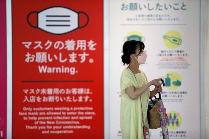 سيدة تمسح مقبض مظلتها وتضع كمامة وتسير أمام توجيهات لمنع انتشار فيروس كورونا في مدخل متجر في شيبويا بطوكيو يوم السبت. تصوير: رويترز.