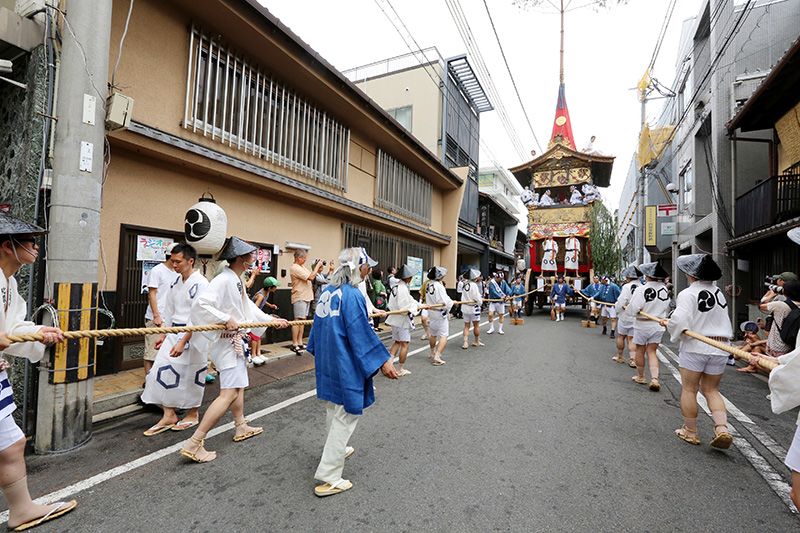 “北观音山”山鉾正行进在新町通街道上。身穿白色短外罩的是“曳方”(用粗麻绳拉山鉾的人),身穿蓝色短外罩的是“车方”(调整彩车方向的人)。因为彩车高度超过20米,所以车顶上也坐了人,以防止彩车撞到电线