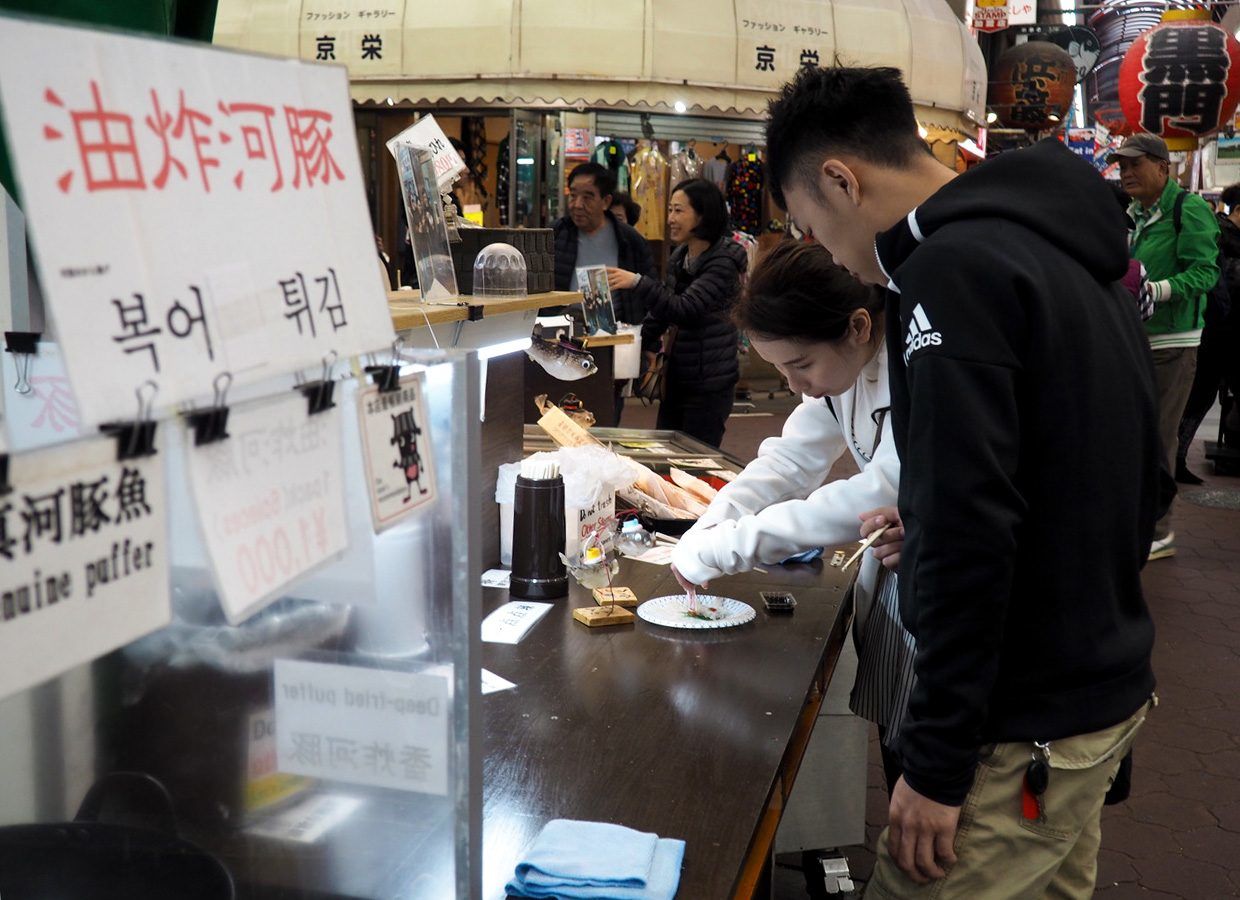 大阪黑门市场的河豚店“mina美”前，站在摊边吃河豚生鱼片的中国情侣（摄影：笔者）