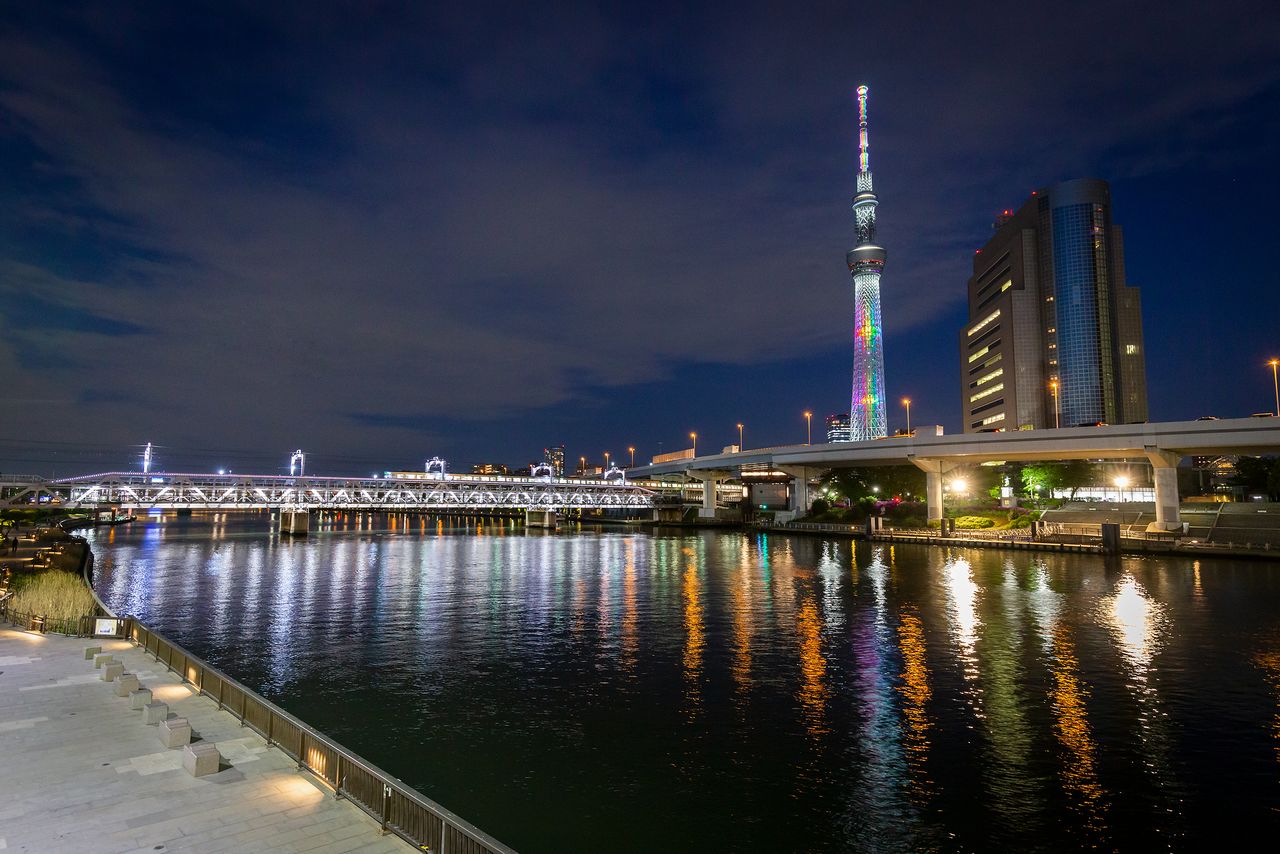 The tower is lit up in modulated shades of white for one hour after sunset. The Sumida Riverwalk, a pedestrian walkway connecting the Skytree area with Asakusa across the Sumida River, runs below the railway bridge span. The structure to the right is the Sumida municipal office.