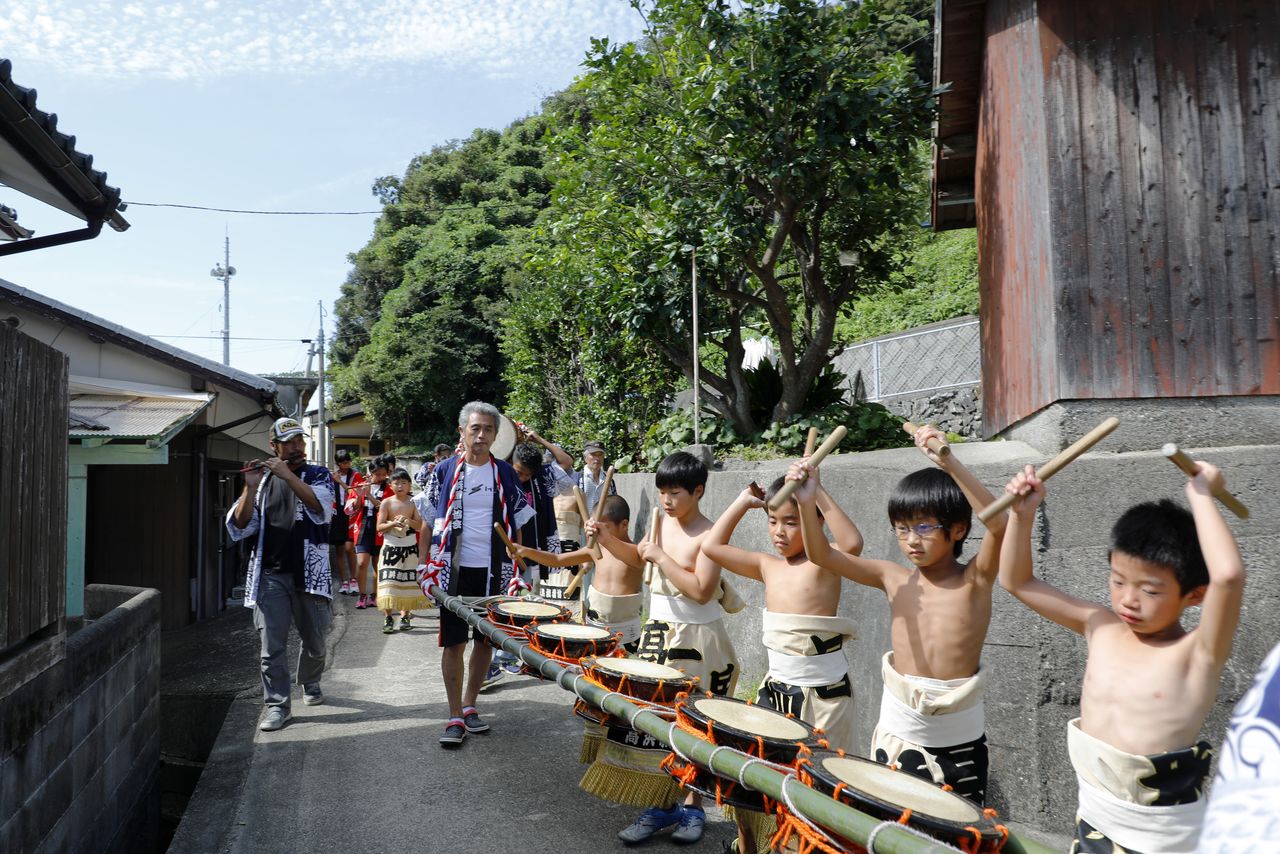 Children in sumō mawashi belts walking in line playing taiko drums attached to green bamboo poles. In northern Kyūshū, they call the autumn festival kunchi. (© Haga Library)