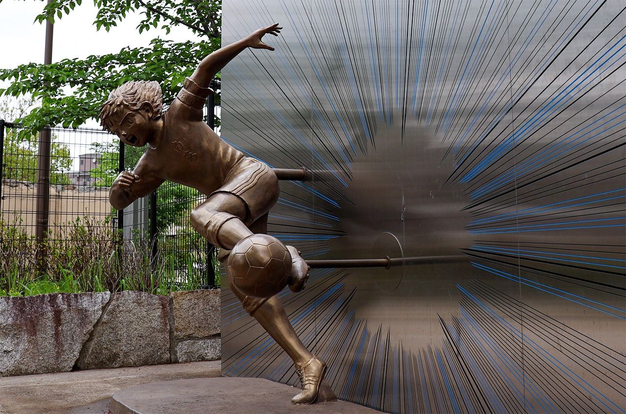 A statue of Ōzora Tsubasa outside the main entrance to Tokyo Metropolitan Minami-Katsushika Senior High School. There is a space where you can pose, with your foot to the ball in place of Misaki Tarō, for a shot with Tsubasa himself. (© Matsumoto Sōichi)