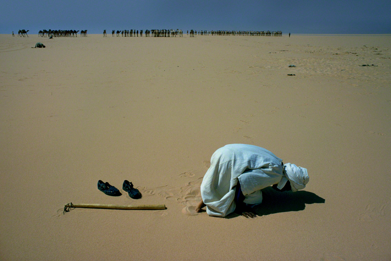 After a simple lunch, a man kneels in the burning sands of the desert to pray in the direction of Mecca. After praying, he turns and returns to join the camel caravan waiting in the background. Photographed in 1975 in Fezzan, in the southwestern interior of Libya. (From Sahara)