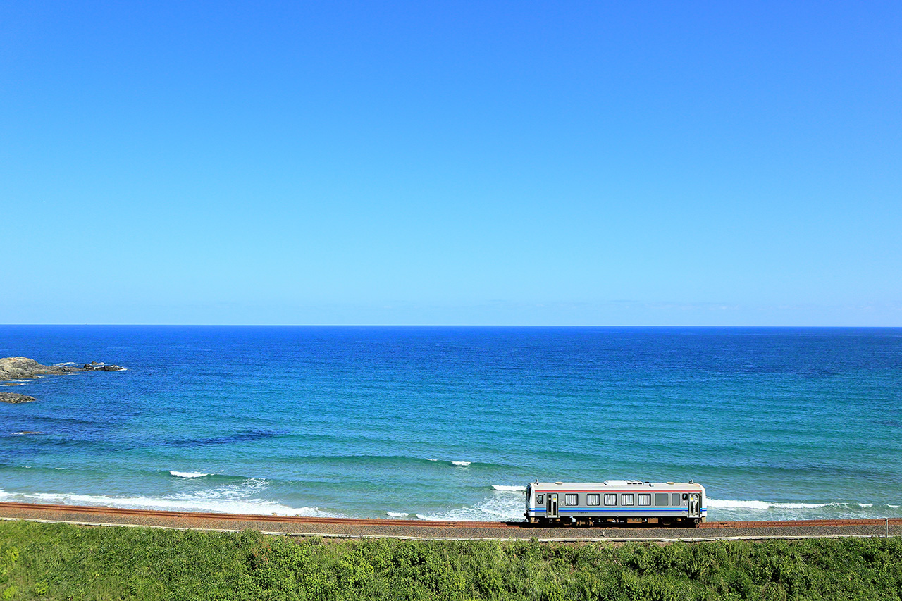 August. A Kiha 120 series on the JR San’in Main Line between Orii and Mihomisumi Stations in Hamada, Shimane Prefecture. There is something cool and refreshing about the way the train trundles along the shore of the Sea of Japan, with its distinctive bright blue hue, under the clear azure sky. It is as if the tracks are floating on the water.