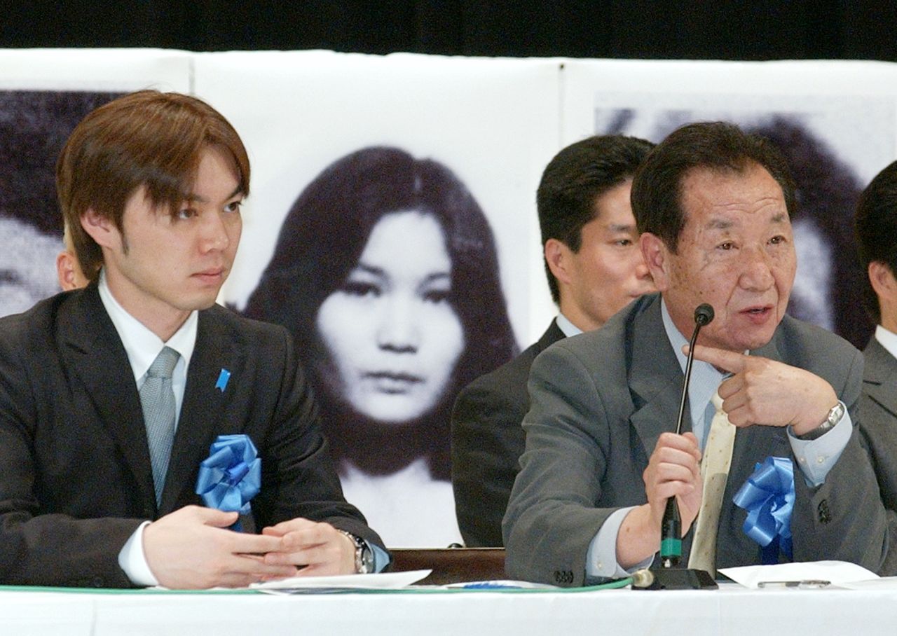 Iizuka Kōichirō (left) was raised as the child of Taguchi Yaeko’s brother Shigeo (right) after his mother was abducted. In the background of this April 2005 photo is an image of Yaeko. (© Jiji)
