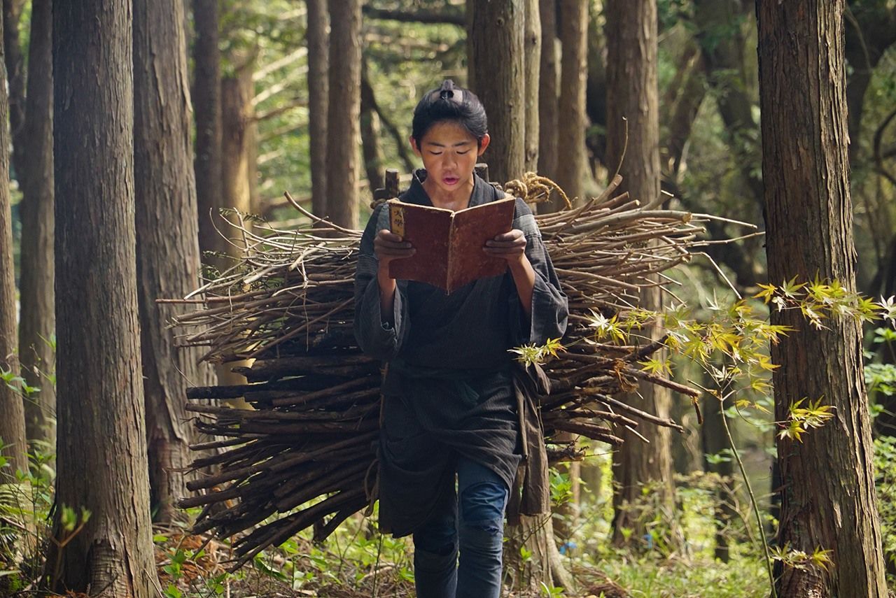 The young Ninomiya (played by Andō Mikoto) steals a moment to study while hauling a load of firewood in the 2019 film Ninomiya Kinjirō. (© Movie Ninomiya Kinjirō Production Committee) 