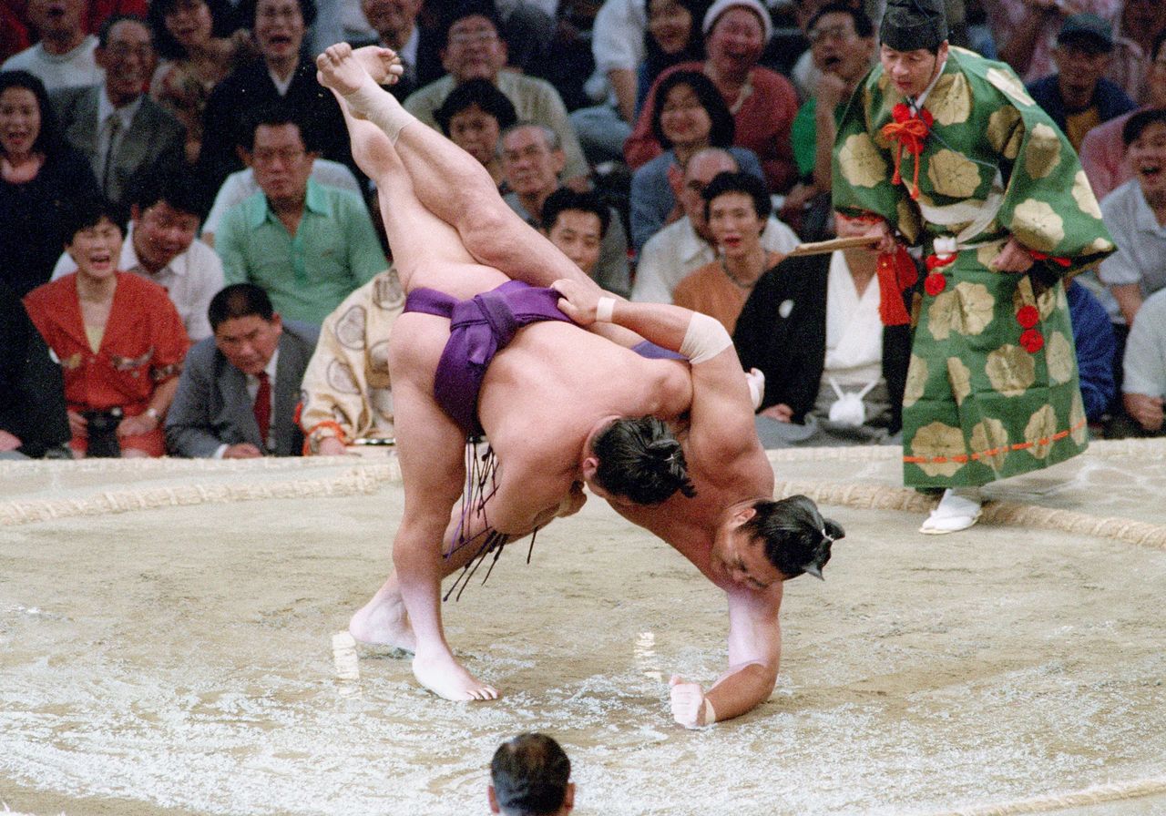 Mainoumi upends ōzeki Kirishima (right) with a spectacular kakenage hooking inner thigh throw. While Mainoumi was known for a broad repertoire of unusual moves, his solid grounding in sumō basics and lower body strength helped him prevail over larger opponents. Taken on July 11, 1993, at the Aichi Prefectural Gymnasium. (© Jiji)
