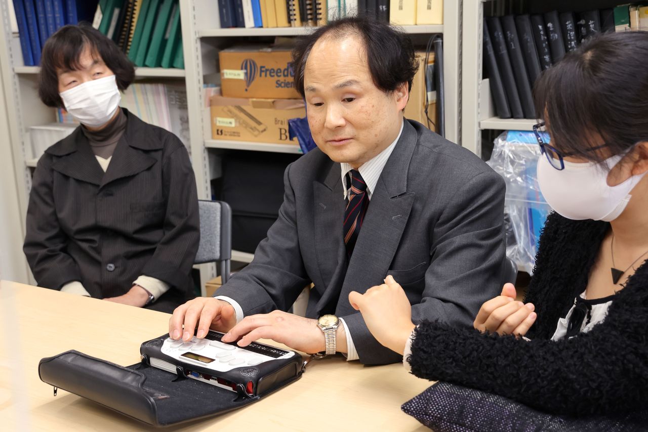 Fukushima demonstrates the use of the BrailleSense mobile device used to input braille and collate information. At left is finger braille interpreter Haruno Momoko. (© Hanai Tomoko)