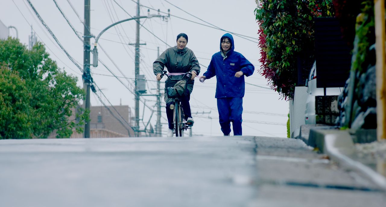 Reiko rides alongside Satoshi, her bicycle tied to his wrist, as he exercises to try to treat his condition. (© Throne/Karavan Pictures)