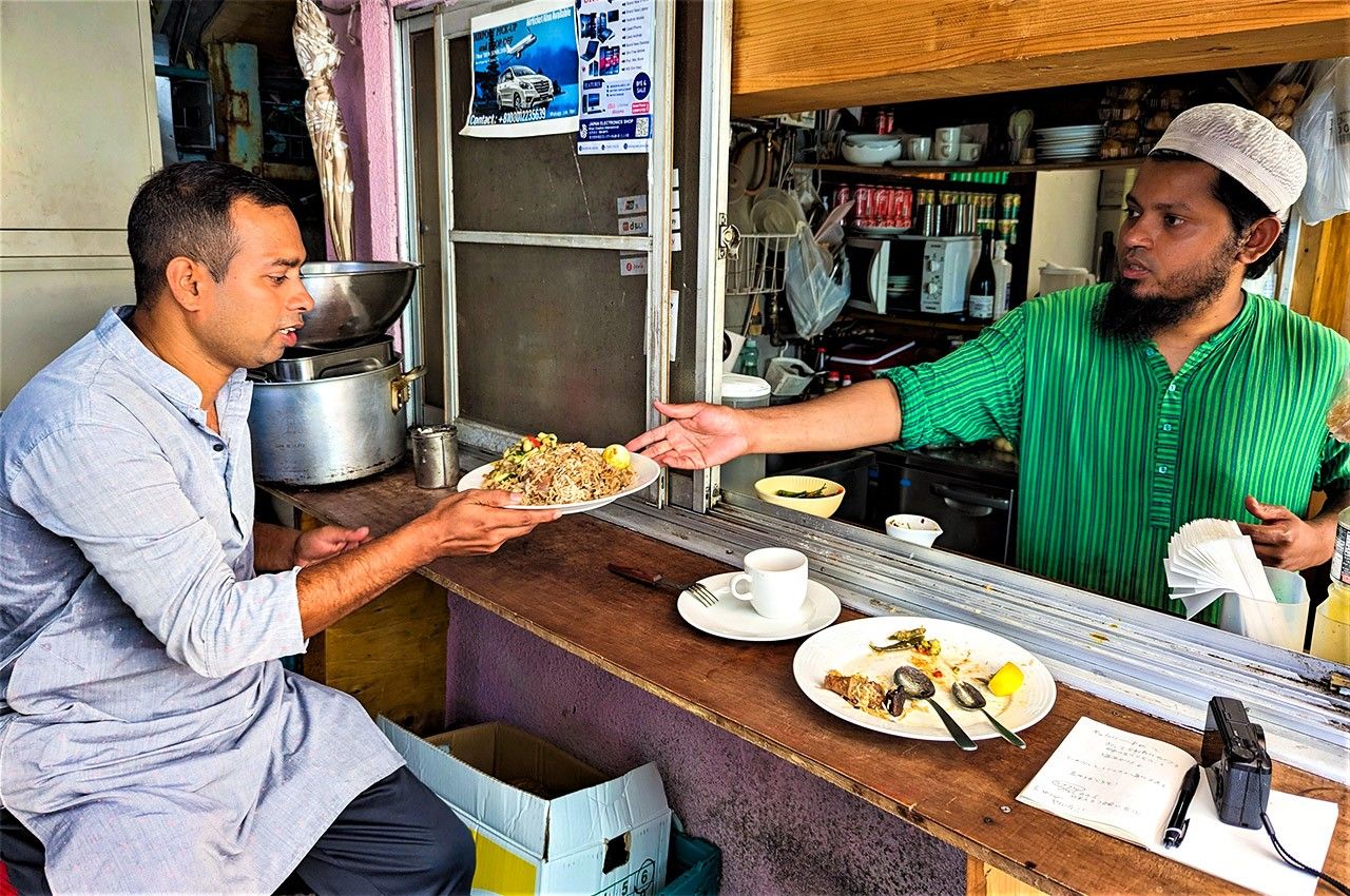 Restaurant owner Ali Jalal serves a biryani to Rana, who is sitting at the sheltered outdoor counter (© Kumazaki Takashi)