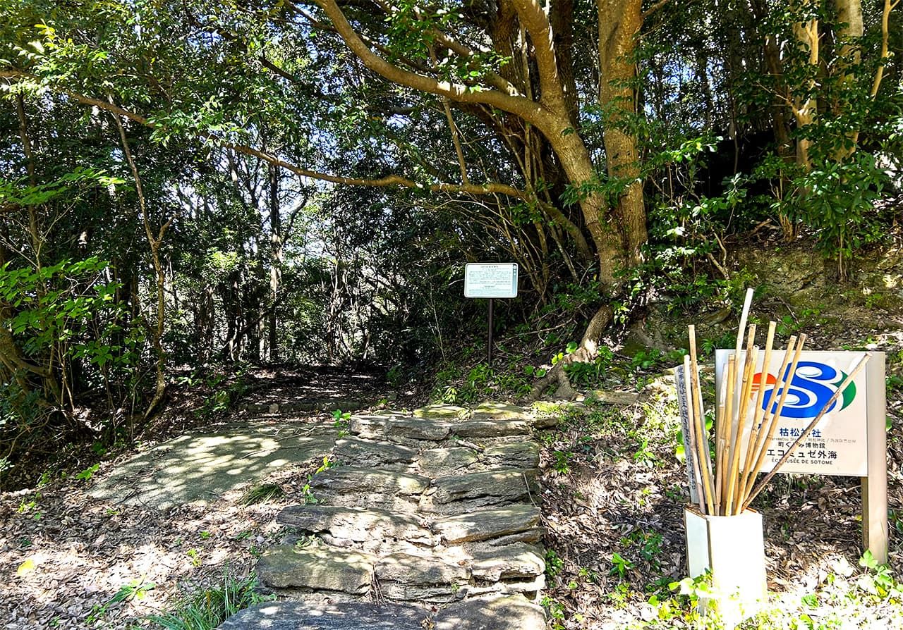 The entrance to Karematsu Jinja. From here, a five-minute walk up a stone stairway leads to the main hall. There are walking sticks for pilgrims in front of the sign. (© Amano Hisaki)