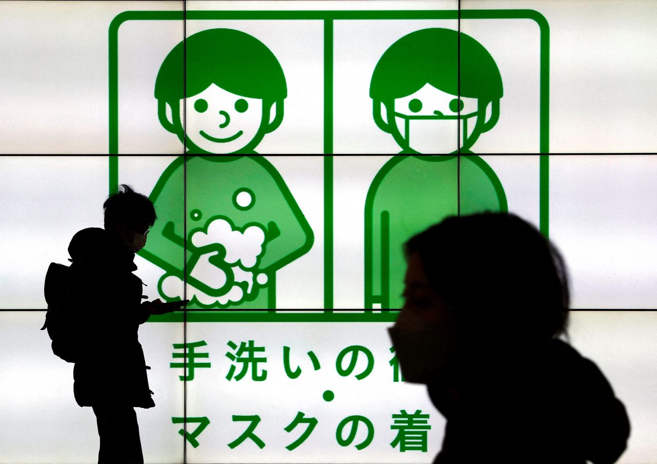 Passersby wearing protective face masks walk past in front of an electric screen displaying notice about COVID-19 safety measures, amid the coronavirus disease (COVID-19) pandemic, in Tokyo, Japan, February 1, 2022. REUTERS/Issei Kato