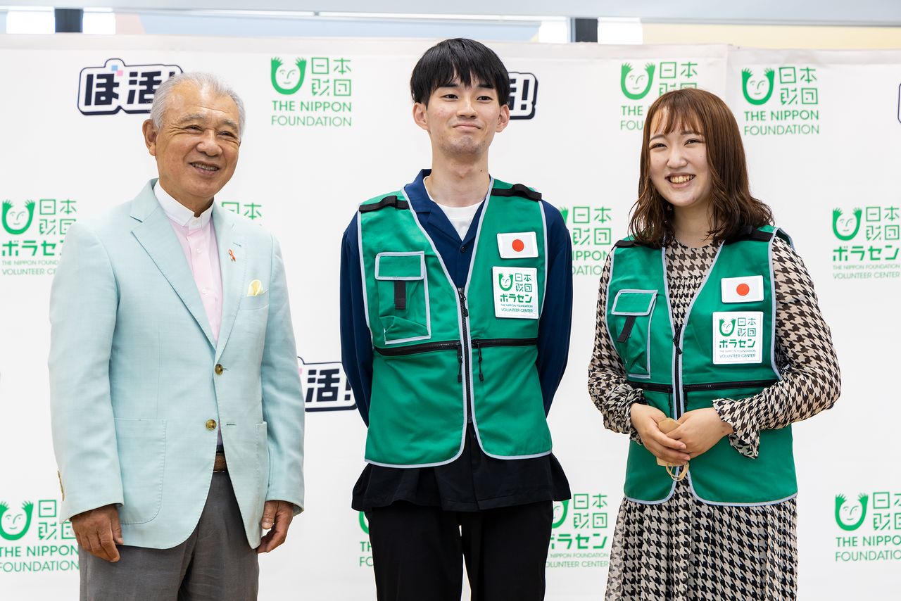 From left, Nippon Foundation Chairman Sasakawa Yōhei poses with Fujita Kōya, a junior at Dōshisha University, and Waseda’s Saitō Rinka, in their volunteer uniforms.