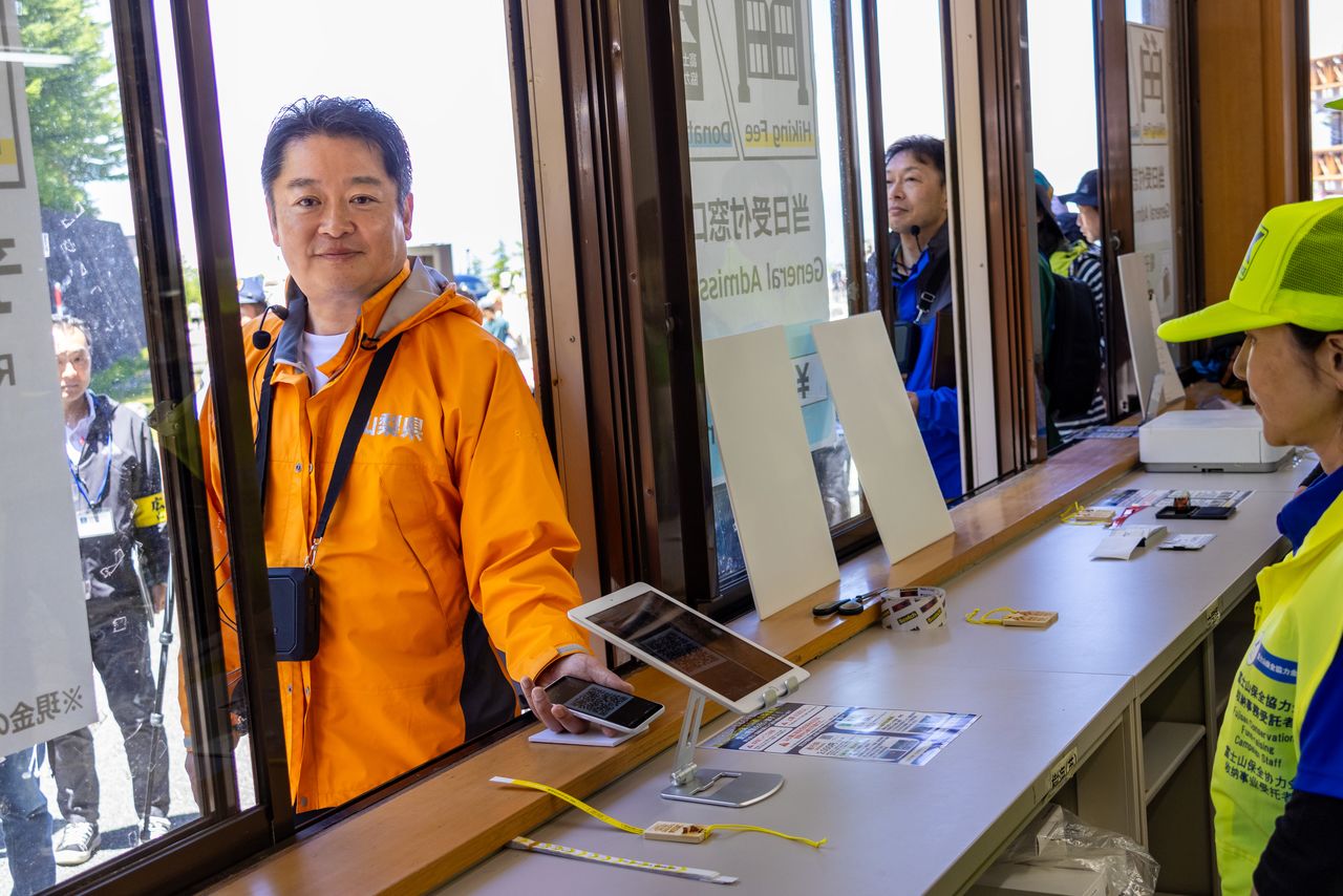 Yamanashi Governor Nagasaki demonstrates how to pass the gate with proof of prepayment on a phone. Hikers are issued a wristband showing they have paid the entry fee. (© Nippon.com)
