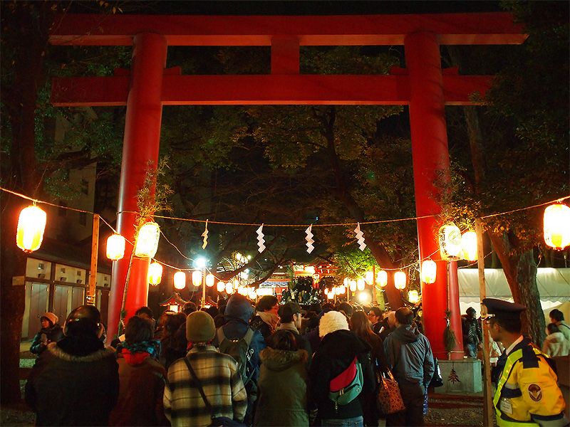 People in jackets and winter hats beneath a Japanese temple arch at night, with round lights on strings.