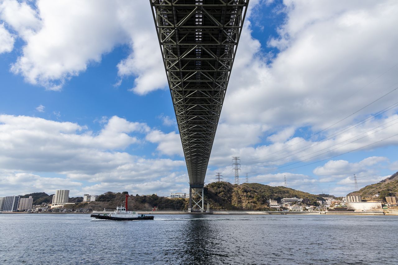Vista de la localidad de Dannoura-chō, al otro lado del estrecho, desde debajo del puente Kanmon, en el lado de Kyūshū. La zona que alberga el grupo de edificios de la derecha es Mimosusogawa-chō, donde se encuentran las ruinas del antiguo campo de batalla de Dannoura y la entrada al lado de Shimonoseki del túnel pedestre Kanmon.
