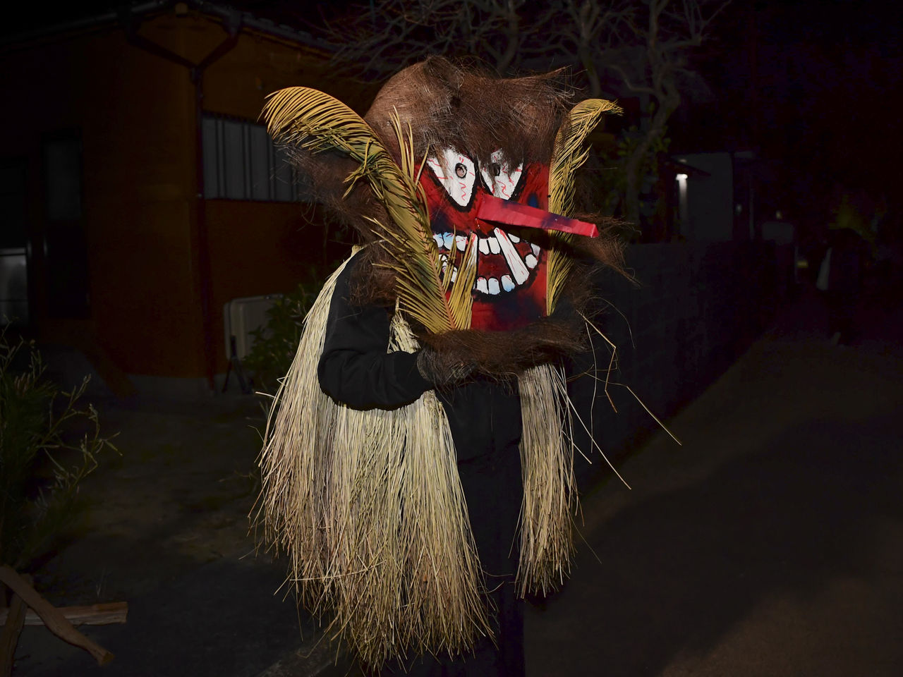 Un toshidon se mueve a través de la oscuridad hacia las casas con niños. El pelo está hecho de corteza de palmera, y los lados de la máscara están adornados con una melena hecha de hojas de palma de sagú.