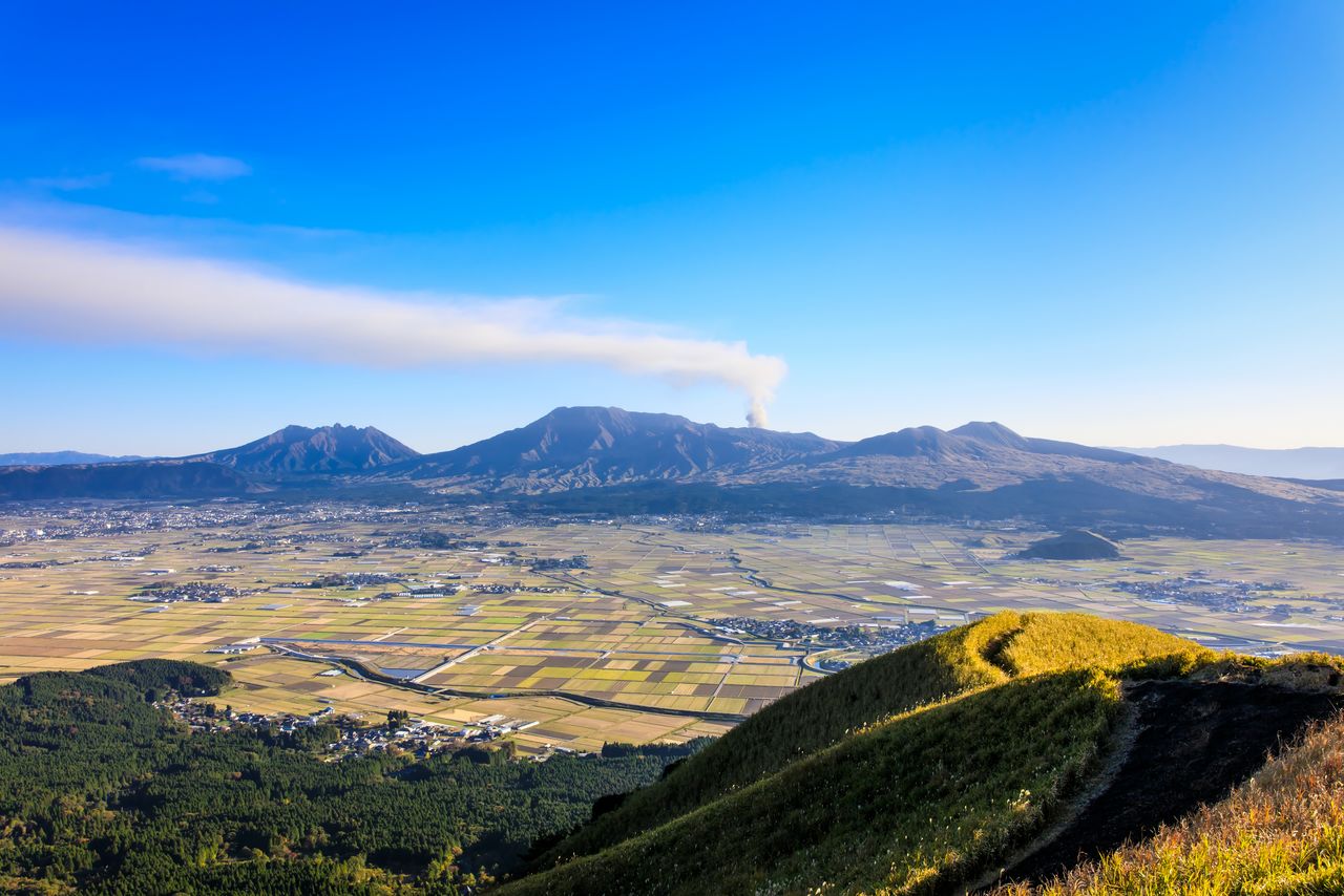 La vista de los arrozales de las calderas y los cinco picos de Aso desde el monte Daikanbō, el borde exterior norte de la caldera. En el fondo, el pico central expulsa una fumarola. Fotografía: PIXTA.