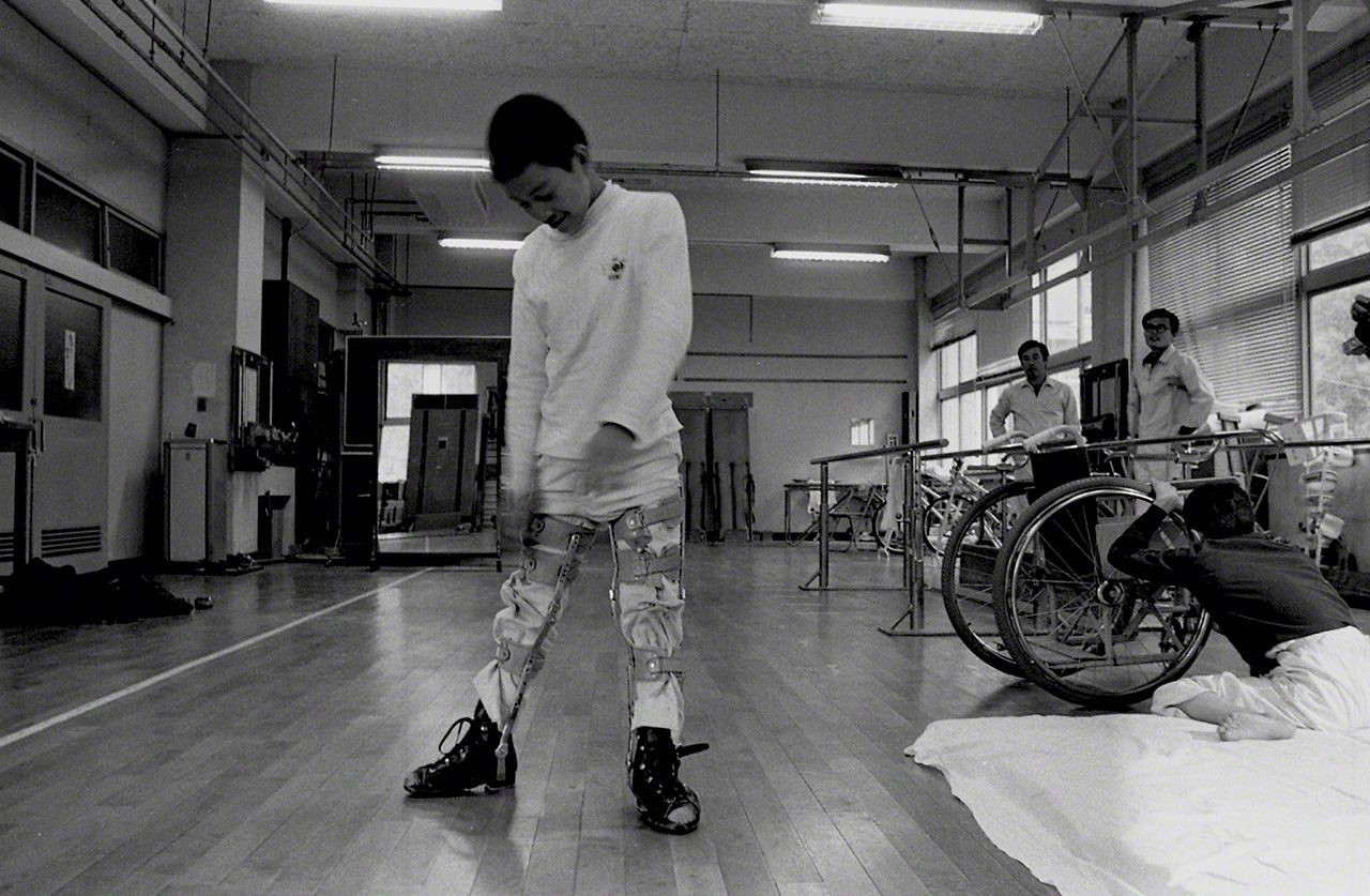 Nagai Isamu (a sus 14 años): Incapaz de caminar desde que era niño, su agenda diaria comprende una dura rehabilitación. No existen tratamientos efectivos. Desde muy joven es un enamorado de las cámaras; en la época de esta fotografía (1973) solía imprimir sus propias imágenes en el estudio de Eugene