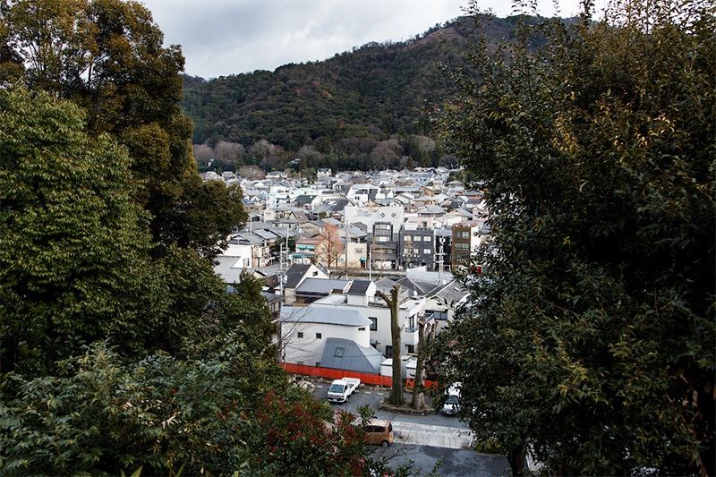 Vista panorámica desde el oeste de la Maison O (diseñada por Nakayama Hideyuki, 2009). El edificio se alza rozando con la valla de color bermellón que rodea el santuario.