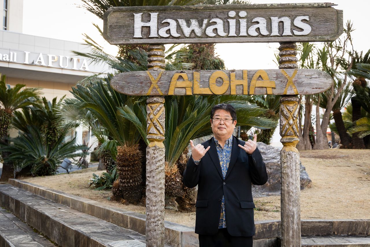 Sekine haciendo el gesto del saludo aloha frente a una palmera.