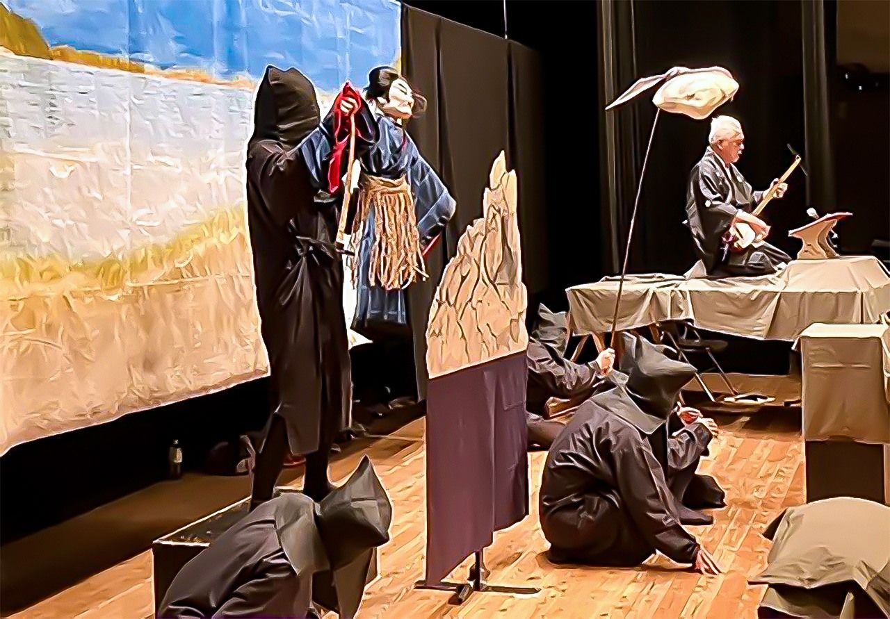 Chlóe, que destaca de incógnito en el centro, lleva la cara tapada y el atuendo negro característico de los marionetistas. Sujeta la marioneta que representa al pirata Kokusen’ya (Coxinga), el personaje histórico que protagoniza la obra Kokusen’ya Kassen, de Chikamatsu Mozaemon. Esta representación tuvo lugar el 19 de octubre de 2024 en Hirado (Nagasaki). A la derecha, el narrador, que toca el shamisen.