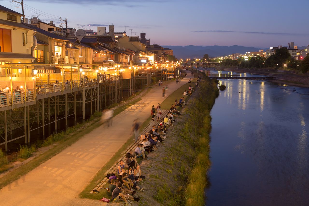 Las terrazas elevadas en las orillas del río Kamo y la gente disfrutando del verano en la ribera. Cerca de 90 restaurantes ofrecen el servicio en las terrazas de mayo a septiembre. (Distrito de Nakagyō, ciudad de Kioto. PIXTA)
