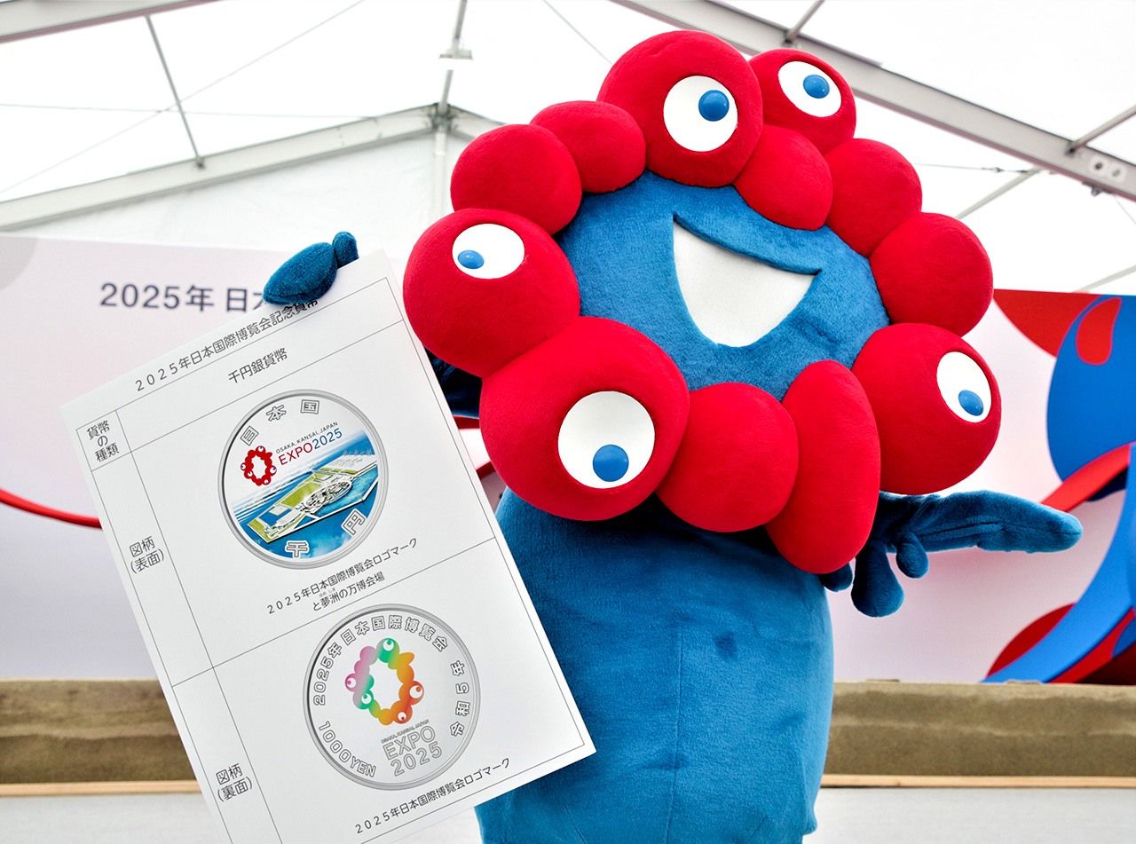 La mascota de la Expo de Osaka 2025 Myaku-Myaku sujeta un cartel con detalles sobre la moneda conmemorativa. (© Jiji)