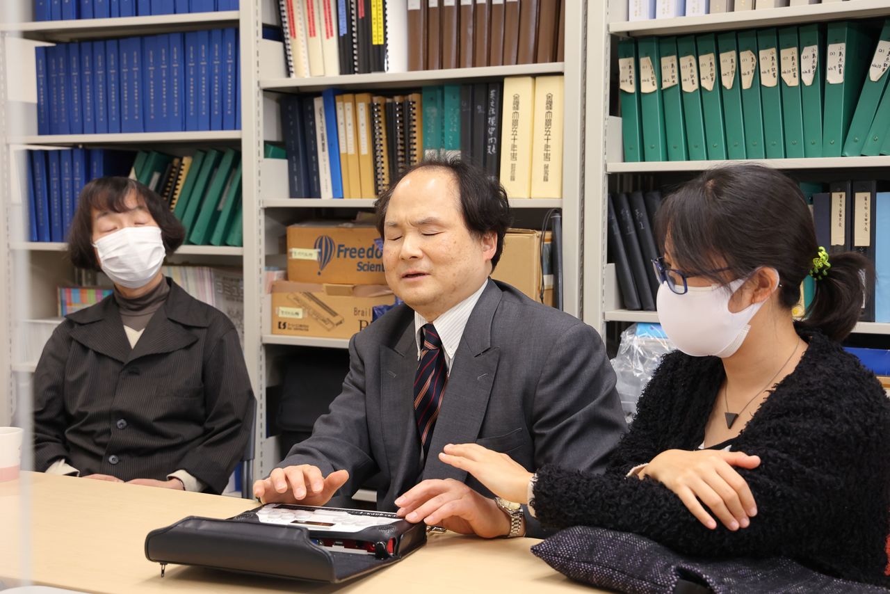 Fukushima muestra el funcionamiento de la terminal BrailleSense leyendo las noticias del día. A la derecha está sentada su otra intérprete, Maeda Atsumi.