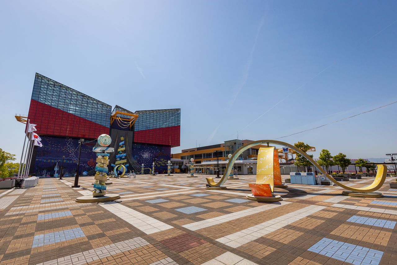 L’aquarium Kaiyûkan d’Osaka vu du côté de la « place du marché » (Market Place), dans le complexe de loisirs et de commerce du Tenpôzan. L’édifice orange que l’on aperçoit sur la droite abrite l’entrée, la billetterie et la boutique des produits vendus exclusivement par l’aquarium. Les objets du premier plan font partie d’un décor temporaire mis en place à l’occasion du 30e anniversaire de sa création.