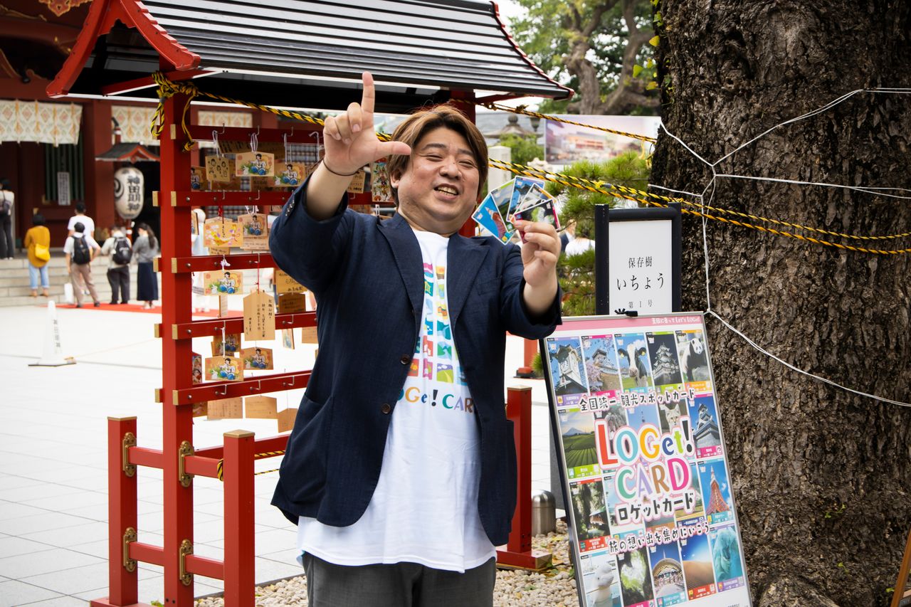 Yamada tient des cartes LOGet devant un des lieux illustrés. Son bref passage en tant qu’employé dans une entreprise de jouets lui a permis de comprendre l’attrait des objets de collection.