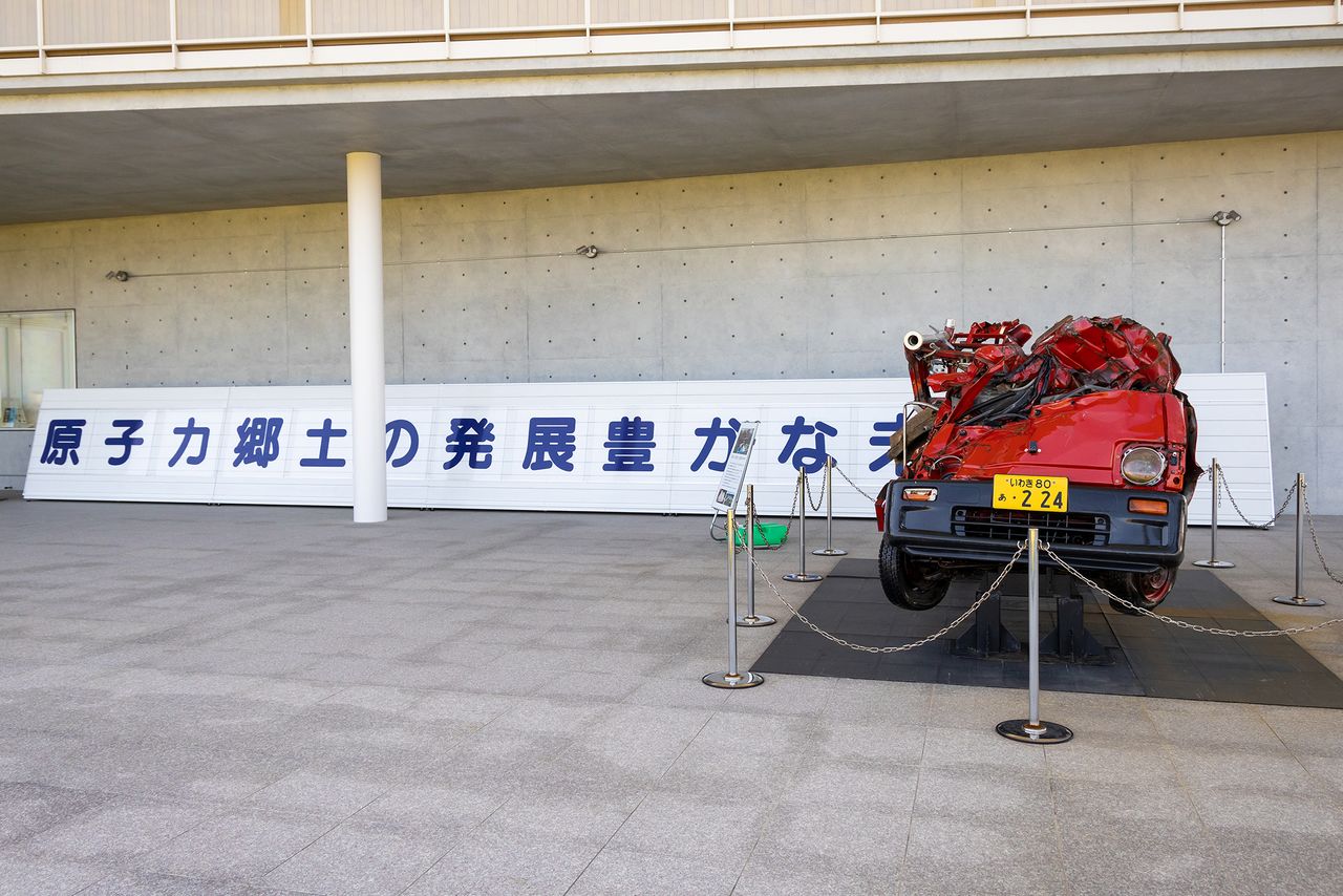 Cette voiture de pompiers, détruite par le tsunami, est exposée à l’extérieur, à côté de la copie d’une banderole de promotion de l’énergie nucléaire qui était autrefois exposée en ville.