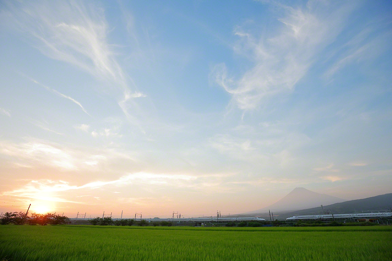 Juillet. Un Shikansen Nozomi de la s&eacute;rie N700A, sur la ligne JR T&ocirc;kaid&ocirc;, entre les gares de Mishima et Shin-Fuji. Les belles perspectives du mont Fuji et des Shinkansen abondent sur cette partie de la ligne. Dans la photo, des Shinkansen allant vers le nord et le sud se rencontrent au pieds de la montagne qui semble flotter dans la brume lors d&rsquo;un beau coucher du soleil.
