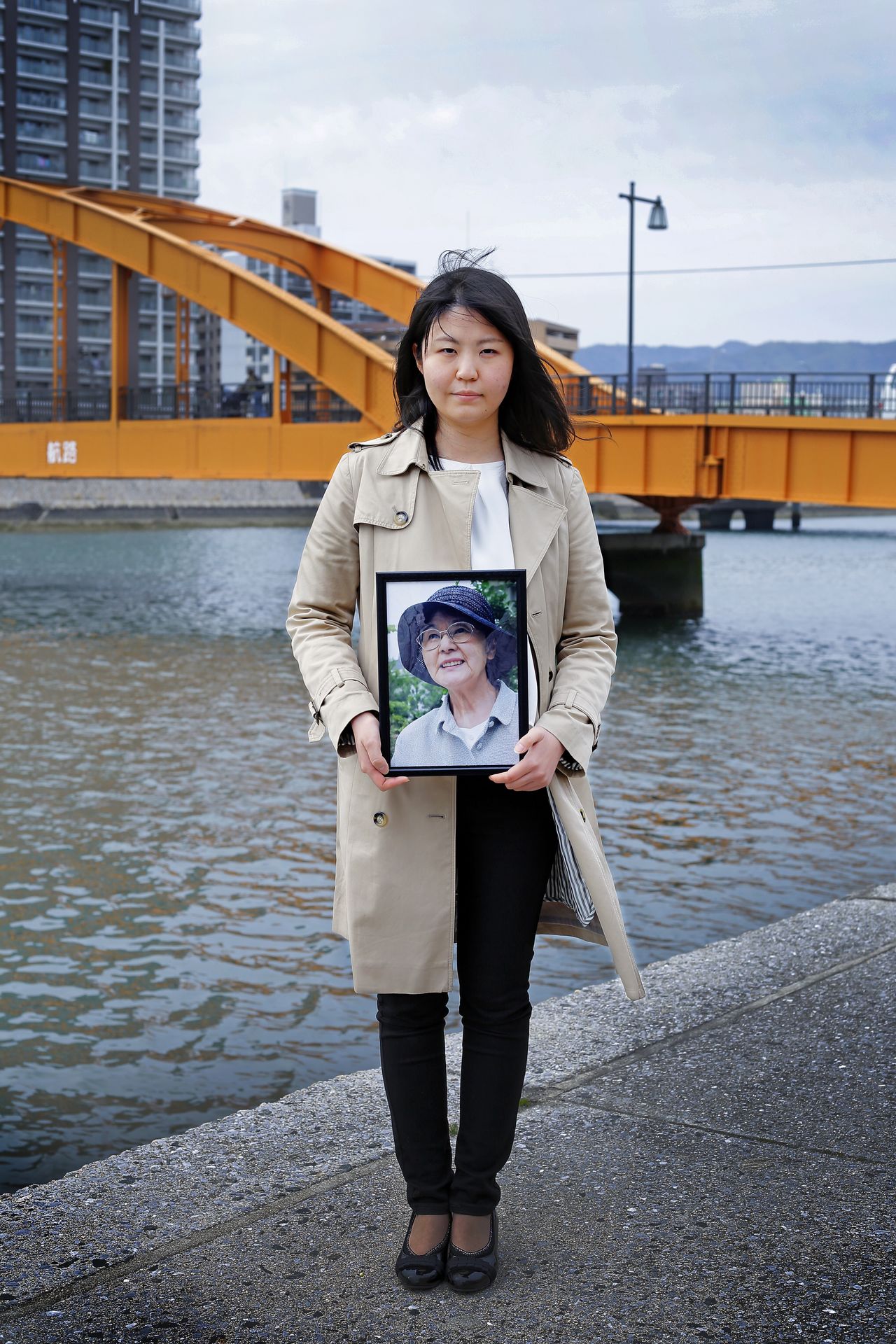 Une participante portant le portrait de sa grand-mère sur le pont Sumiyoshi à Hiroshima, situé à 1,4 km du point d'impact, en mars 2019. Sa grand-mère avait 16 ans et travaillait dans une usine lors du bombardement. Partant à la recherche de sa famille dans le centre-ville, elle a traversé ce pont et a vu d'innombrables corps emportés par la rivière.