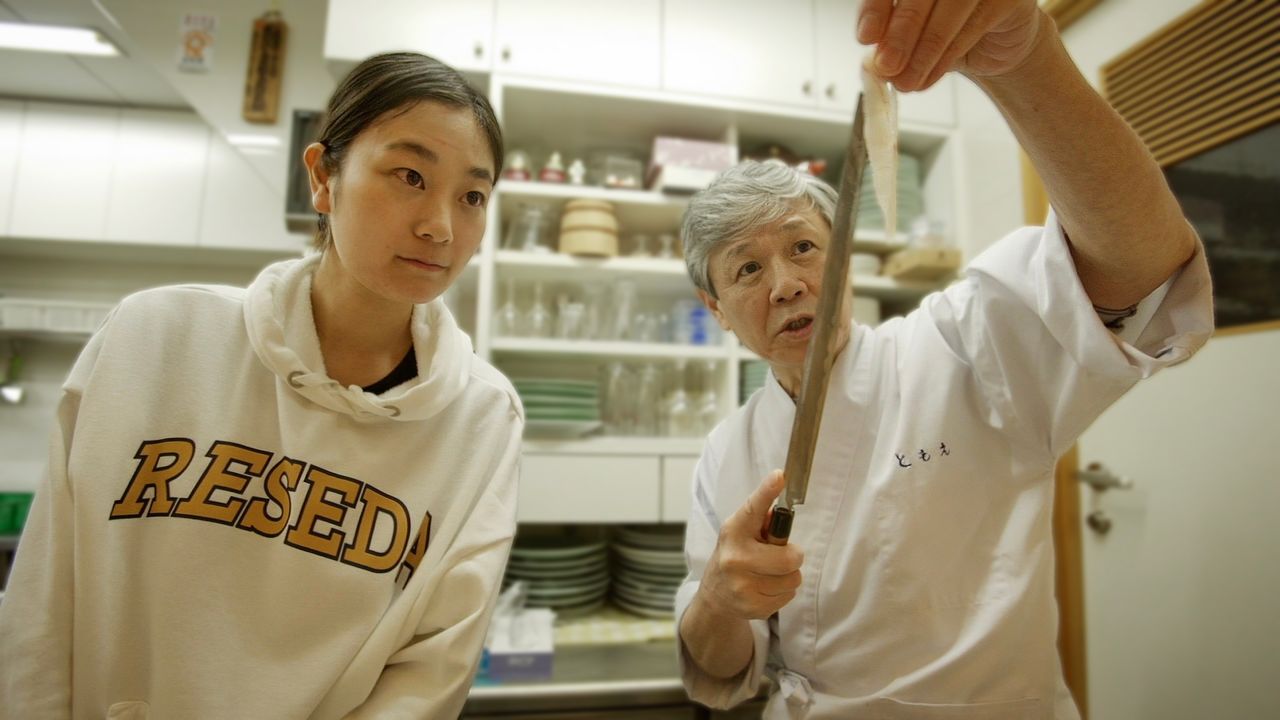 Kamei Kazuhiro (à droite), chef et propriétaire du restaurant « Fugutomoe » à Kyoto, fait une démonstration de la technique de découpe du sashimi de fugu devant Nameki Yukari. (© Gun Fish Film Partners)