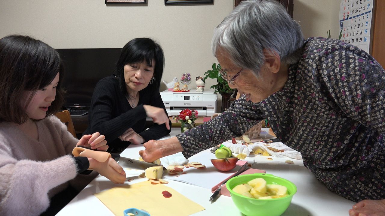 Kimie montre à Moe, qui souffre d’une malformation des mains, comment couper une pomme (extrait du documentaire Gaika) © 2019 SUPERSAURUS