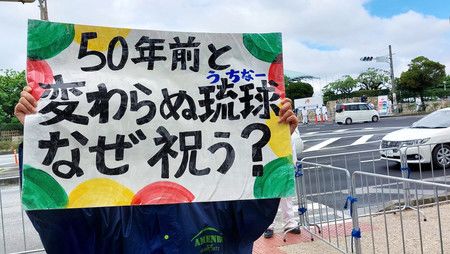 在沖繩回歸50週年記念儀式會場周邊，一位縣民拿起抗議標語＝15日下午、沖繩縣宜野灣市