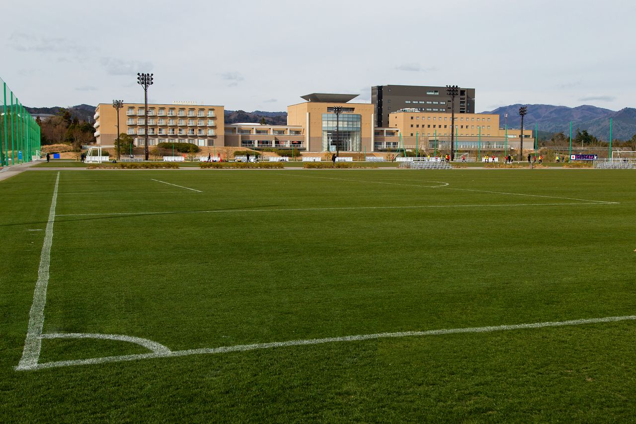 天然芝のピッチ側から、Jヴィレッジの建物群を望む。右奥のグレーの建物が、震災後に新設された宿泊棟「アネックス」