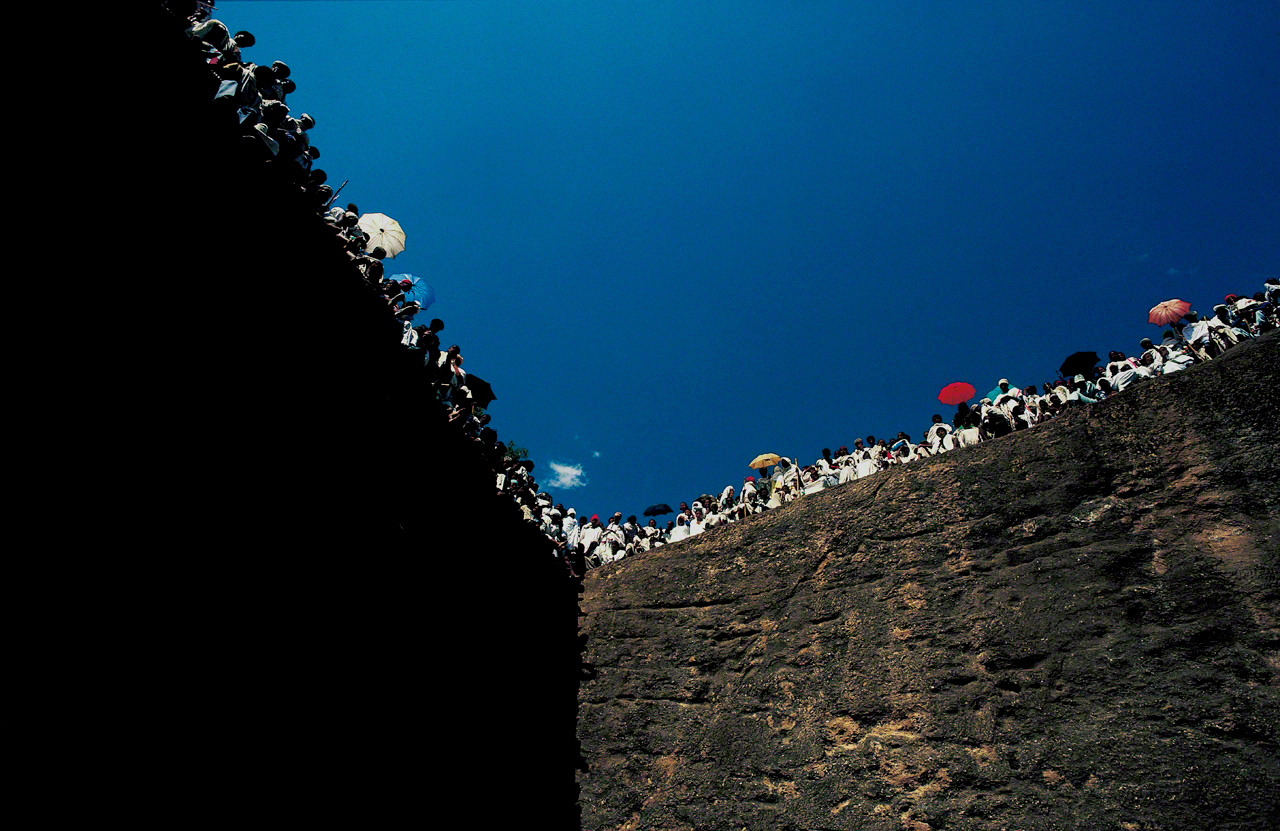 ティムカット（キリスト洗礼の日の祭り）の日、凝灰岩をくりぬいた岩窟教会を取り巻く岩壁に群がり、司祭の説教に聞き入るエチオピア正教会の信徒たち。エチオピア正教は、古代ユダヤ教の流れをくむ、エチオピアで独自に発展したキリスト教。エチオピアのラリベラで1981年撮影。『バハル アフリカが流れる』より