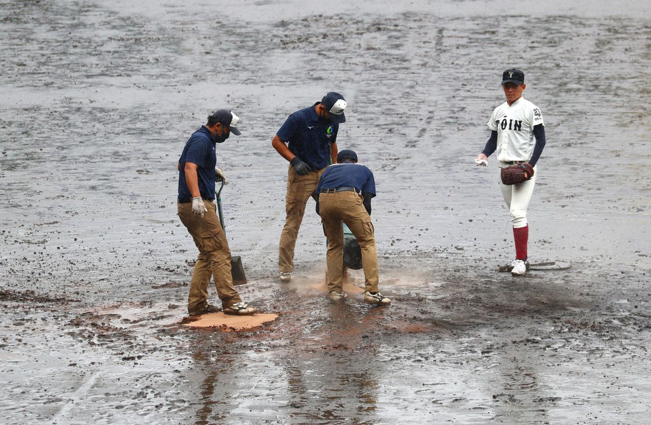 雨の中、グラウンドを整備する阪神園芸のスタッフと、作業を見守る大阪桐蔭高校の竹中勇登投手(2021年8月17日) 時事