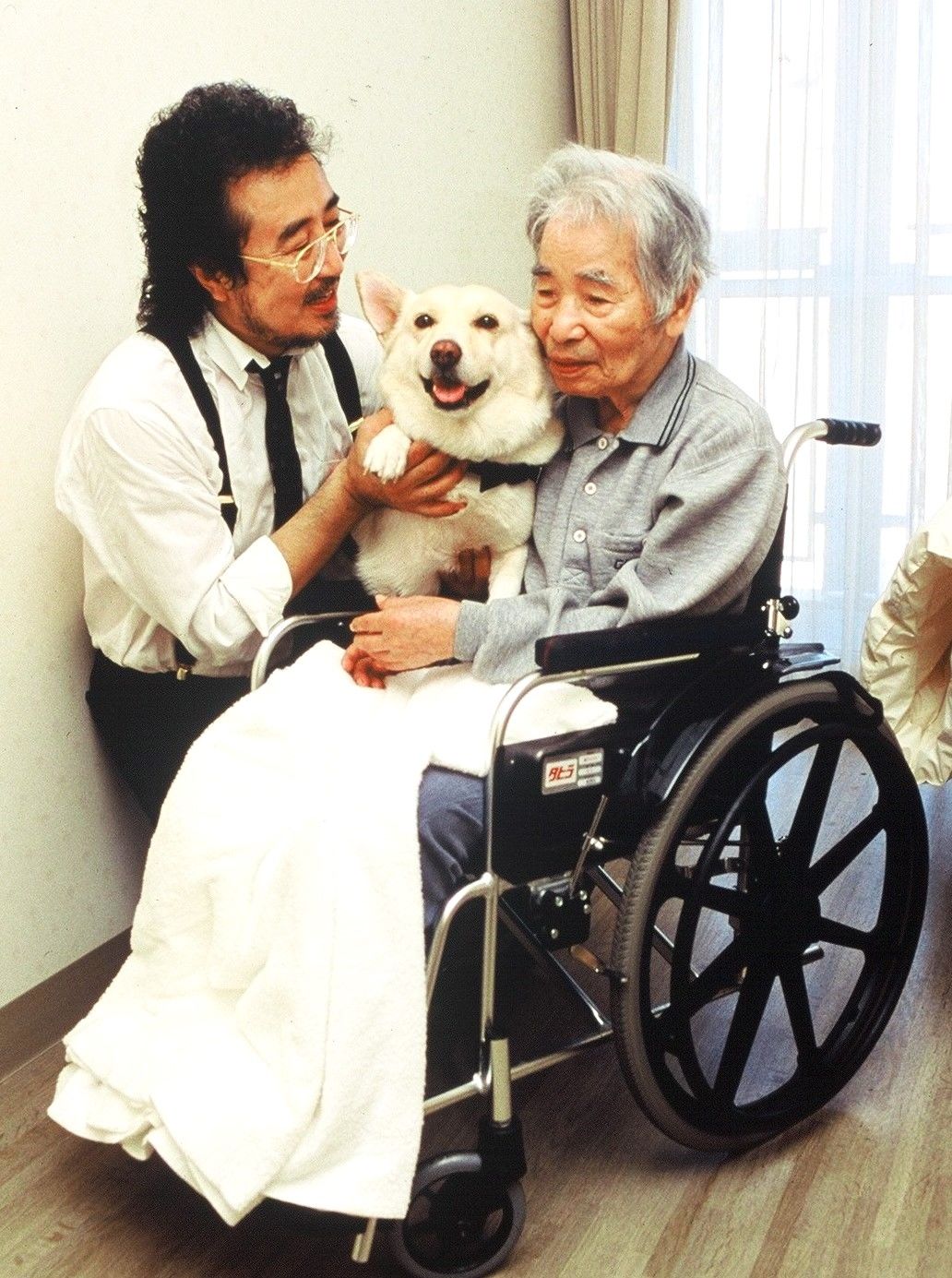 「待っていたよ」とチロリに触れる長谷川外吉さん(右)。チロリに励まされてリハビリに努め、要介護5の状態から自力で立てるまで回復を見せた(左は大木トオルさん) 写真提供=国際セラピードッグ協会