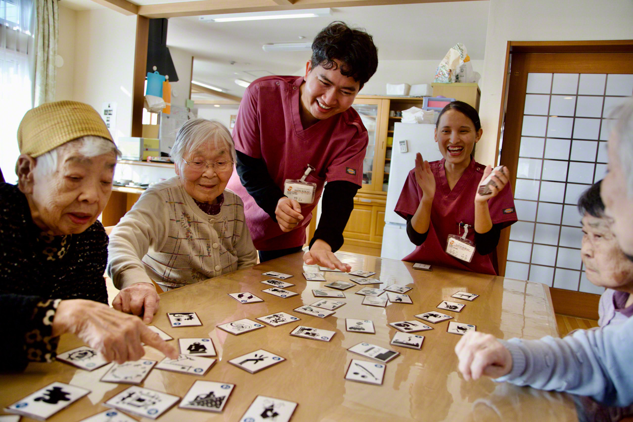 カルタ遊びに興じる。手をたたいて笑うタマンさんと、その横に立つサムさん