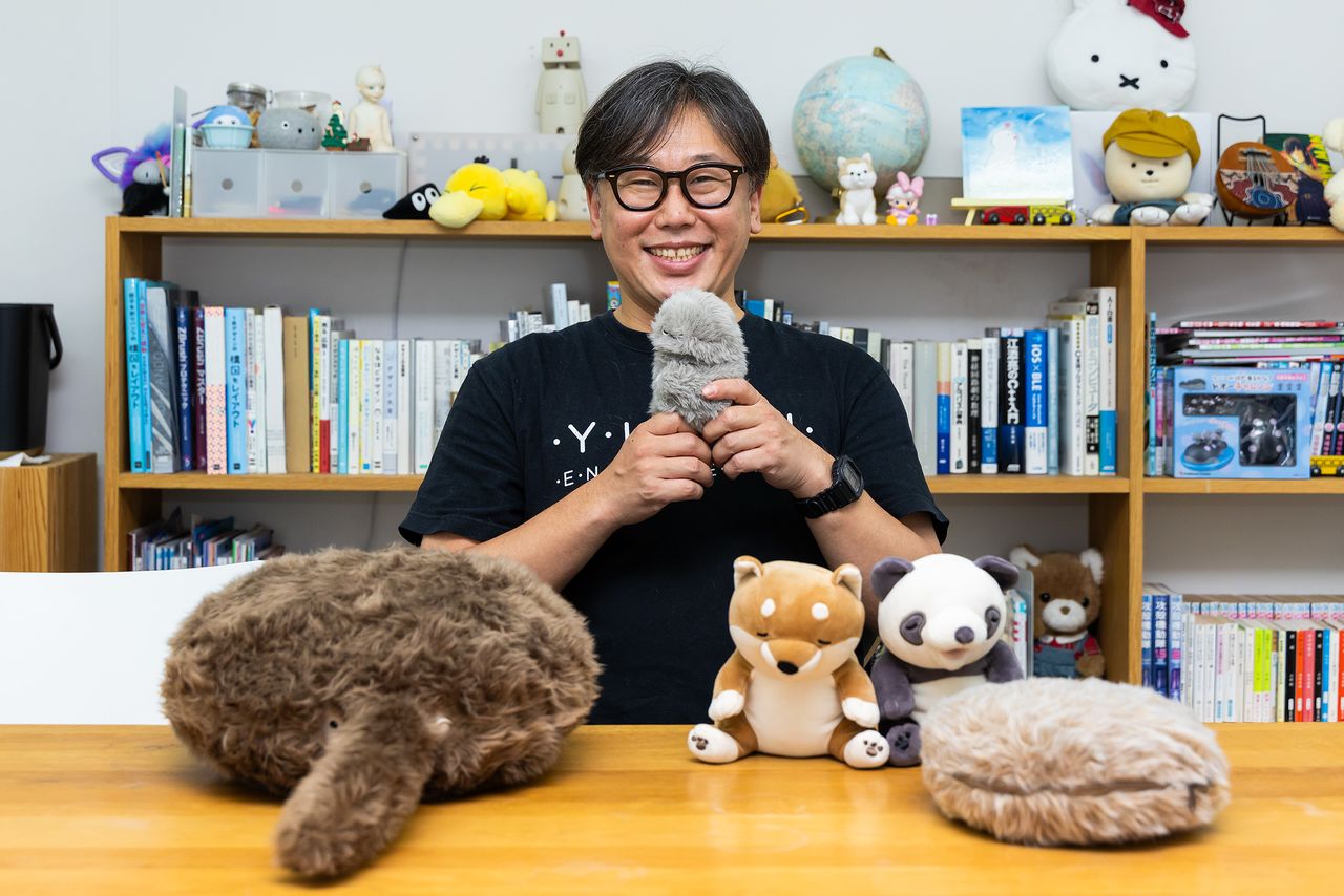 mirumiを抱く青木CEO。テーブル上の左はしっぽのついたクッション型ロボット「Qoobo/クーボ」、右は「PetitQoobo/プチ・クーボ」。なでるとしっぽを振ってこたえてくれる。中央のキツネとパンダは「甘噛みハムハム」(パンダは終売)