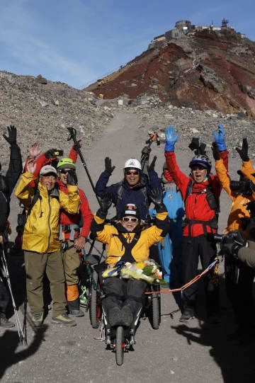 富士山頂に到達し、山岳用車いすに乗りながら万歳する90歳の冒険家三浦雄一郎さん（中央）。後ろは次男の豪太さん＝31日朝