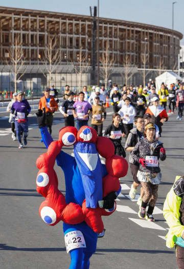 大阪・関西万博の開幕まで100日となり、木造巨大屋根「リング」の前を走るイベントの参加者たち。公式キャラクター「ミャクミャク」に扮(ふん)したランナーもいた=3日午前、大阪市此花区の夢洲